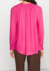 Blusa a maniche lunghe di un rosa brillante, con schiena plissettata, scollatura rotonda e polsini con bottoni, realizzata in un tessuto leggero e testurizzato.