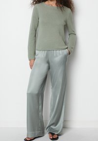 Femme portant un pull en tricot vert clair, un pantalon ample en satin gris pâle, et des sandales noires à bouts ouverts, debout contre un mur blanc.