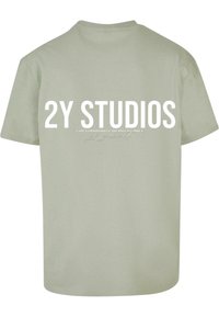 Olīvu zaļš kokvilnas t-krekls ar īsām piedurknēm. Aizmugurē ir liels balts uzraksts "2Y STUDIOS" un smalki akcenta dizaini.