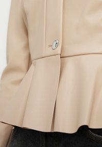 Μπεζ σακάκι peplum από συνθετικό δέρμα, με λεπτομέρειες από γυαλιστερό ασημί κουμπί, μαλακή υφή και πλισέ τελείωμα. Φορεμένο πάνω από σκούρο τζιν.