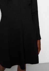 Svart långärmad klänning med en något utsvängd kjol, åtsittande vid midjan, visad på en person mot en ljus och enkel bakgrund.