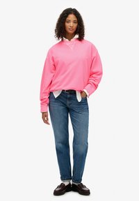 Sudadera rosa de gran tamaño con cuello redondo, superpuesta a una camisa blanca con cuello, combinada con jeans de mezclilla azules y zapatos marrones.
