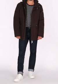Veste marron à capuche avec des fermetures éclair, deux poches avant, portée sur un t-shirt rayé navy et blanc, associée à un jean foncé et des baskets blanches.