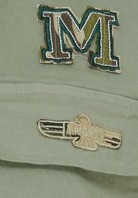 Khaki keps med två broderade märken: ett flerfärgat "M" och en "LUFTFORSEN" örn-design, detaljerad med intrikata sömmar.