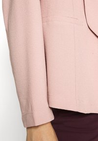 Svetle růžový blazer s texturovanou látkou, s pevně strukturovanými klopami a diskrétní kapsou na levé straně hrudi.