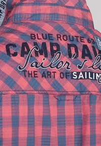 Tissu à carreaux rouge et bleu avec texte brodé. Comprend un motif à carreaux, des coutures contrastantes et une étiquette indiquant "CAMP DAVID".