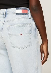 Vaqueros de mezclilla azul claro con un parche en la cinturilla que presenta colores rojo, blanco y azul. Incluye un pequeño logo en el bolsillo trasero.