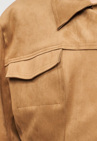 Καμία μπεζ γούνα με μυτερό γιακά, μεγάλο τσέπη στο στήθος και εμφανείς ραφές. Η υφή είναι απαλή και λεία, ενισχύοντας τη συνολική σχεδίαση.