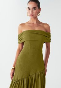 Olivegrünes schulterfreies Kleid aus strukturiertem Stoff, enganliegende Brustpartie und ausgestellter, gestufter Rock. Goldene Creolen und ein klobiges Armband setzen Akzente.