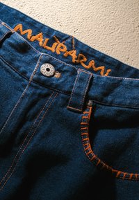 Jeans in denim blu scuro con dettagli di cuciture arancioni, dotati di chiusura con bottoni e logo ricamato "MALÌPARMI" sulla vita.