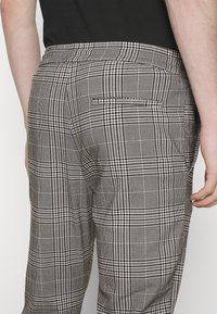 Grijs en zwart geruite broek met een achterzak, een getailleerde pasvorm en een gladde stofstructuur. Zijaanzicht met zicht op de tailleband en hand.