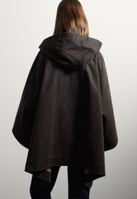 Poncho à capuche marron foncé avec une coupe décontractée, présentant un ourlet arrondi et des détails de couture visibles. Le matériau semble texturé et durable.