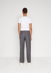 Femme portant une chemise blanche à manches courtes, un pantalon gris droit et des chaussures blanches, debout face à un fond uni gris clair.