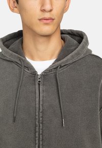 Sudadera con cremallera en gris, hecha de un tejido suave, que cuenta con capucha con cordones, cremallera frontal con tirador metálico y forro interior en contraste.