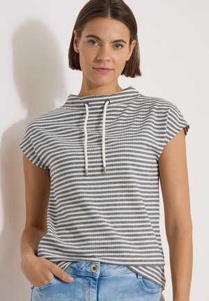 Jeune femme aux cheveux courts et bruns portant un haut sans manches rayé gris et blanc avec des cordons et un jean bleu clair.