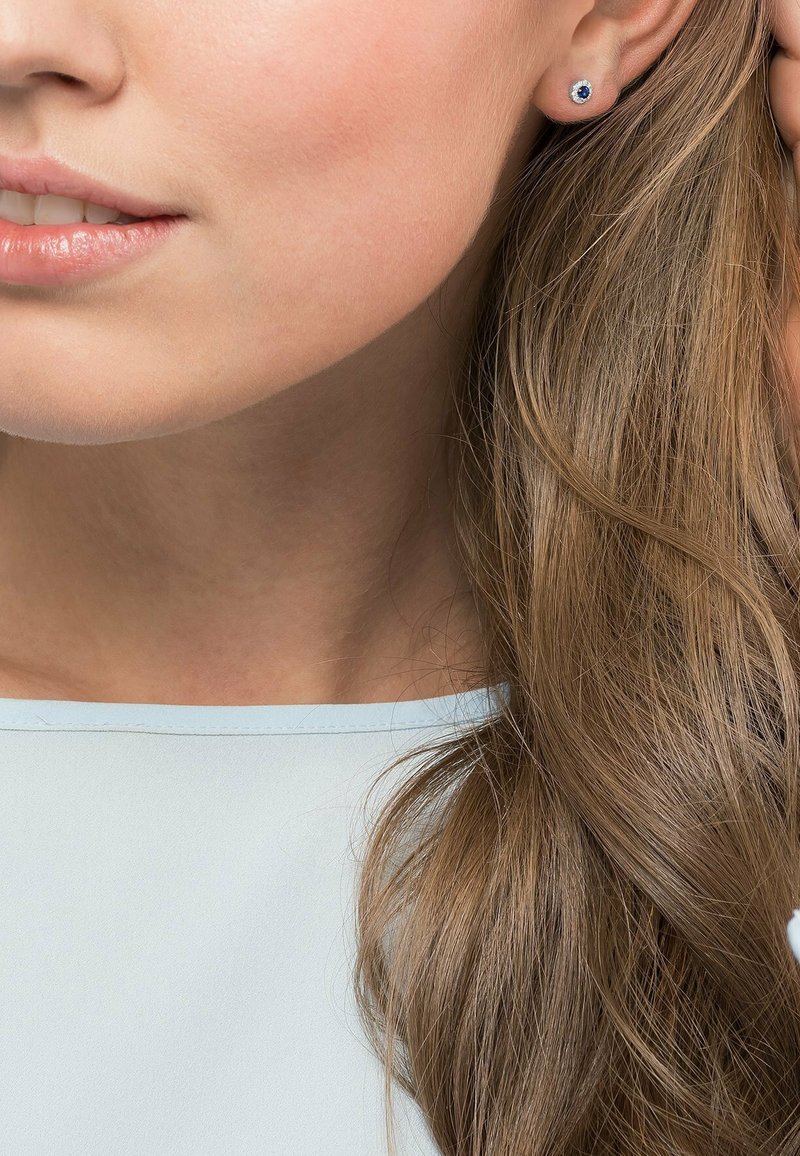 Kleine Ohrstecker mit einem runden blauen Edelstein, umgeben von einer silberfarbenen Fassung, getragen von einer Person mit welligem braunem Haar.