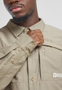 Licht kaki shirt met een kraag en een knoopsluiting aan de voorkant, met een logo, een gladde textuur en zichtbare stikseldetails op de mouwen en de placket.