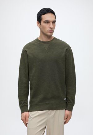 Mann mit kurzen schwarzen Haaren, der einen dunkelgrünen Pullover-Sweatshirt und beige Hosen trägt, steht vor einem einfarbigen hellen Hintergrund.