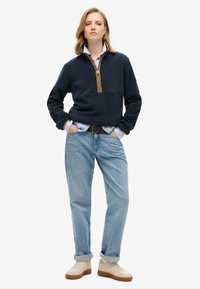 Donkerblauwe fleece pullover met een kwartrits en beige ritsdetails, gecombineerd met lichtblauwe jeans en beige sneakers met bruine zolen.