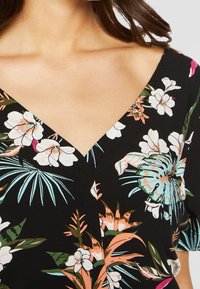 Mujer que lleva un vestido negro de cuello en V con un patrón floral colorido que presenta hojas y flores tropicales en verde, blanco, rosa y naranja.