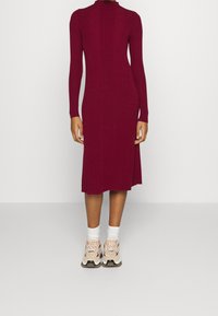 Robe longue en tricot bordeaux à manches longues avec un col haut et une texture côtelée, présentant une longueur mi-mollet et une silhouette ajustée, associée à des baskets beiges.
