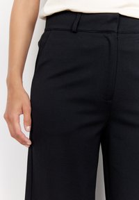 Pantalons ajustés noirs en tissu lisse et légèrement texturé. Présentent un devant plat, des poches latérales et une taille mi-haute.
