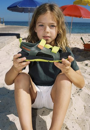 Barn sidder på en sandstrand og holder en grøn og gul Havaianas sandal med parasoller og hav i baggrunden.