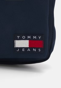 Borsa di tessuto blu navy con un pannello rettangolare con la scritta "TOMMY" sopra una striscia bianca e un quadrato rosso, e "JEANS" sotto, tutto ricamato.