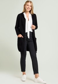 Cardigan noir à capuche, avec poches avant et ouverture sur le devant, porté sur un chemisier blanc et un jean noir, assorti à des baskets blanches.