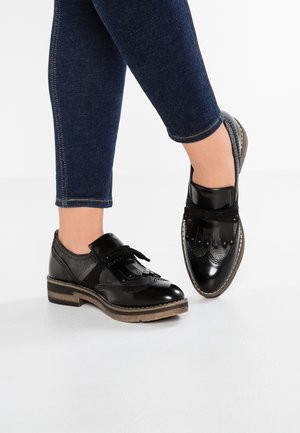 Fødder iført sorte læder brogues med frynse detaljer og snørebånd, parret med mørkeblå tætsiddende jeans på en hvid baggrund.