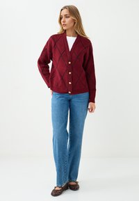 Kobieta stoi w czerwonym kardiganie zapinanym na guziki z czarnym wzorem w romby, w niebieskich dżinsach i brązowych plecionych balerinach na białym tle.