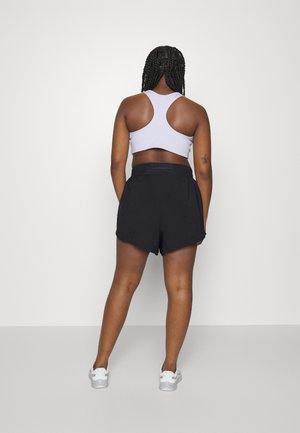 Kobieta z zaplecionymi włosami, ubrana w biały biustonosz sportowy, czarne spodenki i białe trampki, stoi twarzą do gładkiej białej ściany z rękami w kieszeniach.