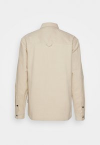 Cream-colored long-sleeve shirt with a button-down collar, textured fabric, single back yoke, and buttoned cuffs. Features a loop at the neck.