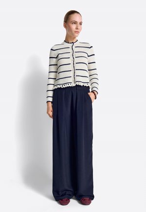 Naine, kes kannab valget ja meresinist triibulist kardigani, laiade jalgadega meresiniseid pükse, burgundia värvi jalanõusid ja pruuni käevõru, seisab ühe käega taskus.