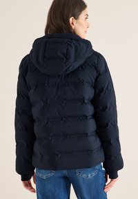 Kvinde iført en marineblå quiltet hættejakke og blå jeans, stående med ryggen til mod en ensfarvet lys baggrund.