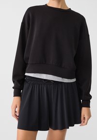 Sweat-shirt noir court avec un col rond et des épaules tombantes, superposé à une chemise grise. Associé à une jupe noire à taille ajustable par cordon.