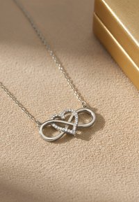 Collana d'argento con simbolo dell'infinito intrecciato a un cuore, decorata con cristalli trasparenti. Su uno sfondo chiaro e strutturato.