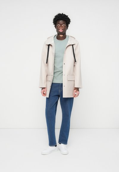 Beige regnfrakk med hette, svarte snorer, to frontlommer, over en lys grønn skjorte, kombinert med blå jeans og hvite joggesko.
