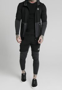 Zwart en grijs sportkleding bestaande uit een jas met rits en capuchon, een aansluitend topje en taps toelopende broek, gecombineerd met zwarte sneakers.