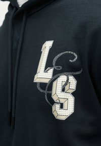 Κοντινή λήψη της περιοχής στο στήθος μιας μαύρης κουκούλας με μπεζ και λευκά υφασμάτινα γράμματα "L" και "S" που διαπλέκονται με ένα διακοσμητικό γκρι φιόγκo.