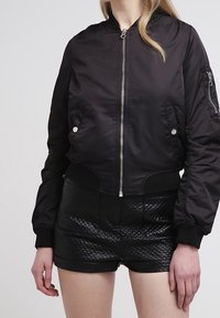 Svart bomberjacka med dragkedja fram, ribbade ärmslut och krage; har silverfärgade detaljer och en sidoficka med dragkedja. Bärs över svarta vadderade shorts.