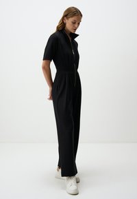 Tuta nera con maniche corte, chiusura con zip, gambe affusolate e tasche laterali. Indossata con sneakers bianche, dal design semplice.