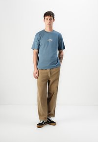 Ung man som står mot en vit vägg, iförd en blå logotyp-t-shirt, beige byxor och svarta sneakers med vita snörningar, med tatuering på vänster arm.