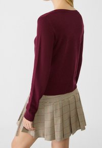 Bordeauxkleurige gebreide trui met lange mouwen, gecombineerd met een beige geruite geplooide rok met rode en zwarte accenten en een gestructureerde stof.