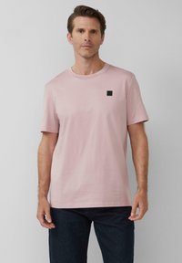 Helles rosa T-Shirt mit kurzen Ärmeln aus Baumwolle, mit rundem Halsausschnitt und einem kleinen schwarzen Logo-Patch auf der oberen linken Brust.