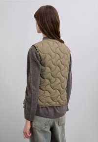 Olive green quilted vest with a textured pattern, worn over a gray long-sleeve shirt, featuring a simple cut and no visible fastenings.
