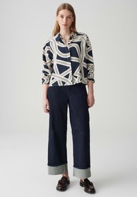 Camicia button-up blu navy con motivo astratto bianco; abbinata a pantaloni denim scuri a gamba larga con orlo in denim chiaro. Mocassini marroni.