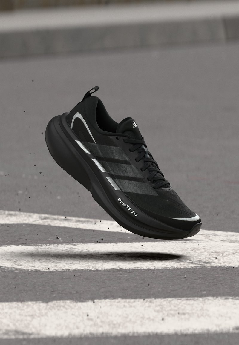 Μαύρο αθλητικό παπούτσι Adidas με αντανακλαστικές ρίγες και τη λέξη "DREAMSTRIKE GLOW" στο πλάι, ελαφρώς στον αέρα πάνω από άσφαλτο με λευκές σημαδέματα.
