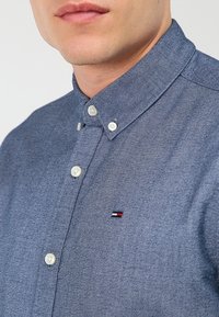 Blå knapp-up skjorta i texturerad tyg, med klassisk krage och vita knappar. Inkluderar en liten logotyp med röd, vit och blå.