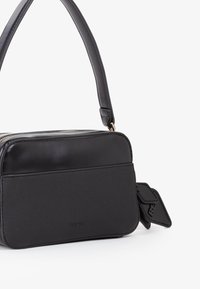 Borsa a tracolla rettangolare nera con pannello frontale testurizzato, parte superiore liscia, manico corto e piccolo etichetta attaccata su sfondo bianco.